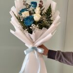 Exquisite bouquet with 3 white roses, 2 blue roses, and seasonal fillers wrapped in white paper with a blue ribbon